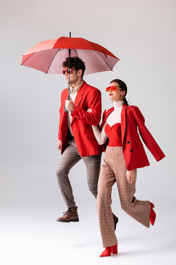 volledige weergave van trendy paar in rode blazers en zonnebrillen met paraplu op grijs - Foto, afbeelding