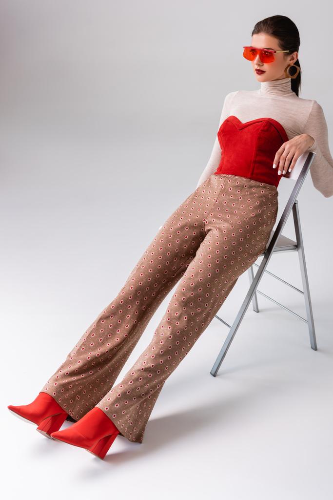 Stilvolle Frau sieht weg, sitzt auf Stuhl mit verlängerten Beinen auf grau - Foto, Bild