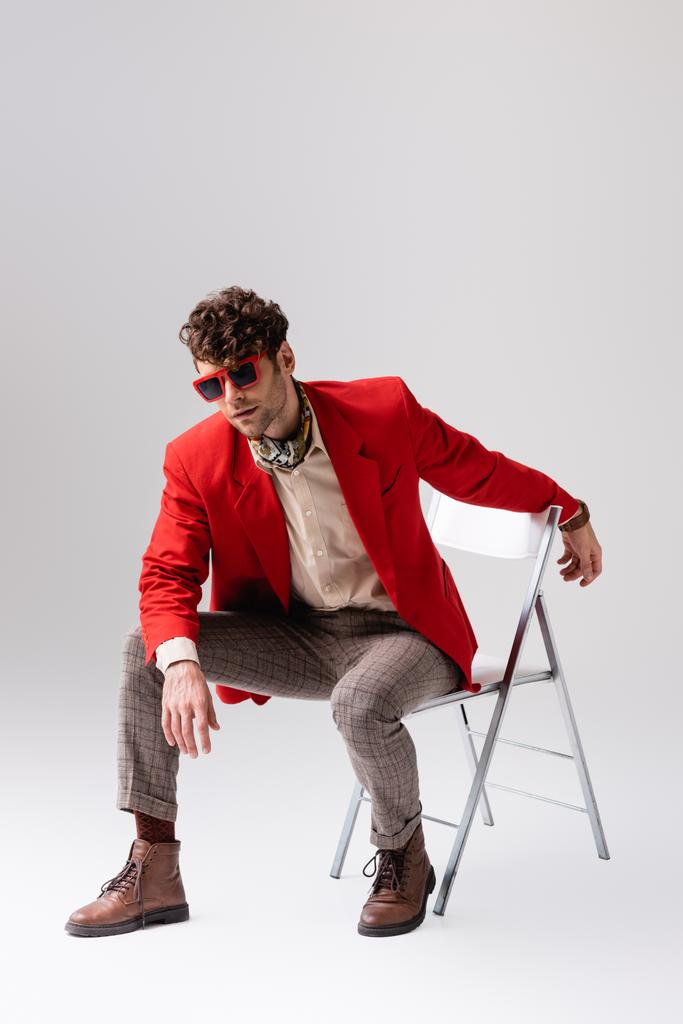 Elegante uomo in blazer rosso appoggiato in avanti e in posa mentre seduto sulla sedia sul grigio - Foto, immagini