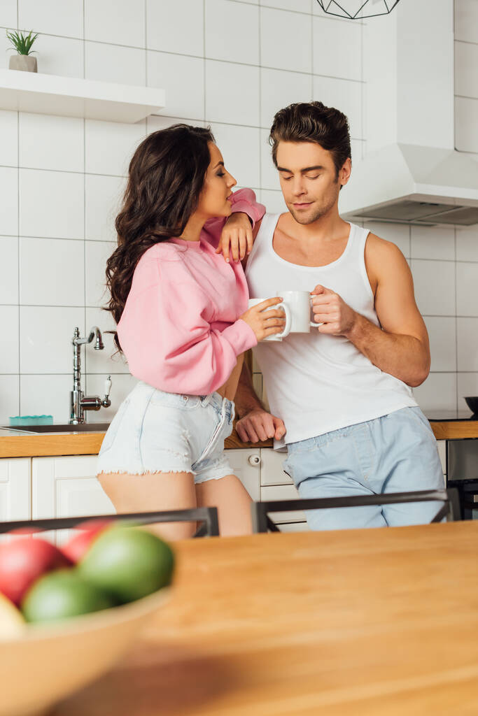 Selective focus of young couple with cups of coffee standing near worktop in kitchen  - Photo, Image