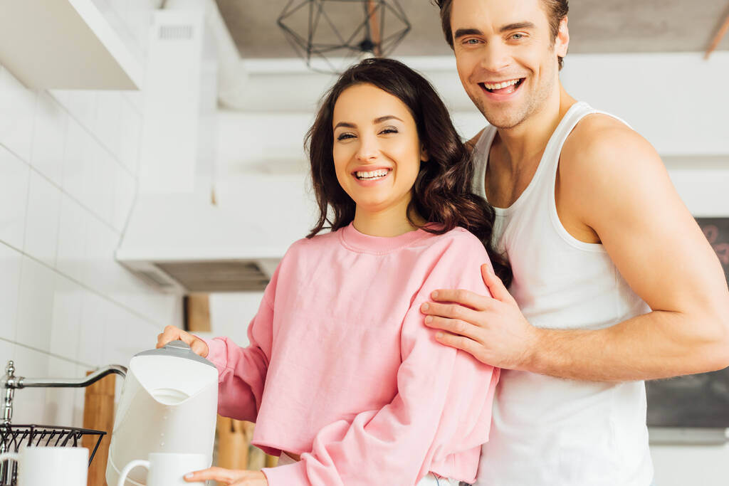Yakışıklı bir adam mutfaktaki çaydanlığı ve fincanı tutan neşeli kız arkadaşını kucaklıyor.   - Fotoğraf, Görsel