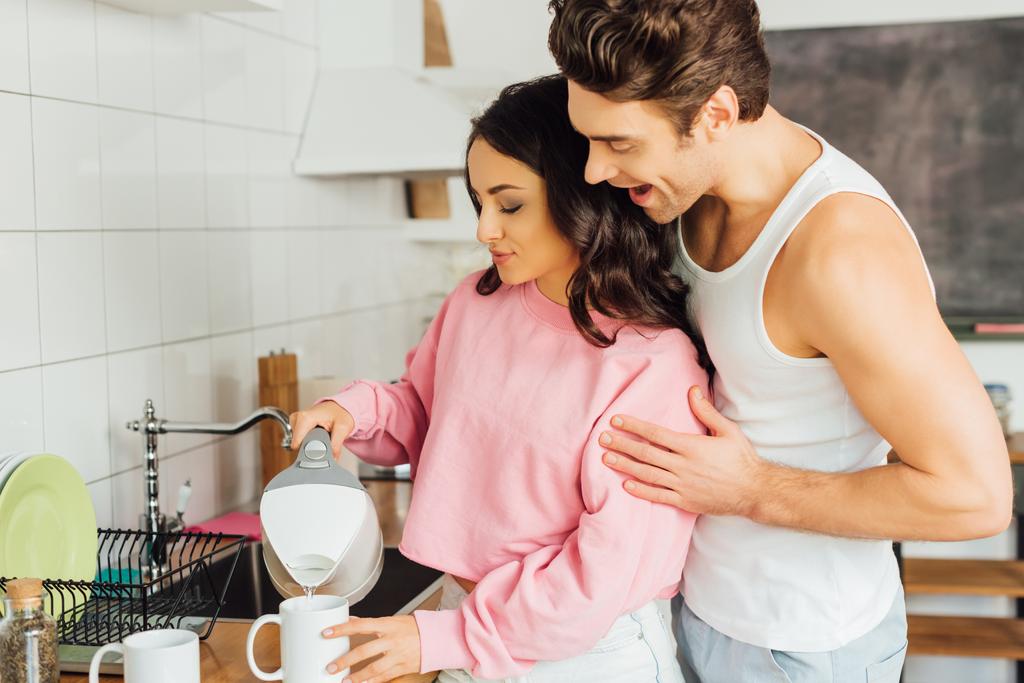 Позитивный мужчина обнимает привлекательную женщину наливая воду из чайника в чашку на кухонный стол  - Фото, изображение