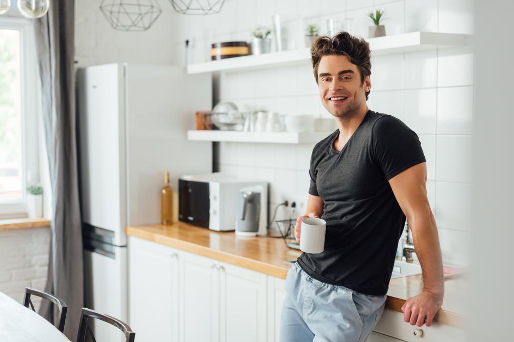 Επιλεκτική εστίαση του όμορφου άνδρα χαμογελώντας στην κάμερα, ενώ κρατώντας ένα φλιτζάνι καφέ κοντά στην επιφάνεια εργασίας στην κουζίνα  - Φωτογραφία, εικόνα