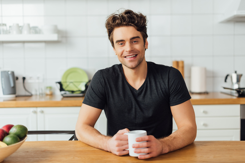 Enfoque selectivo del joven sonriendo a la cámara mientras sostiene la taza de café en la mesa en la cocina
  - Foto, Imagen