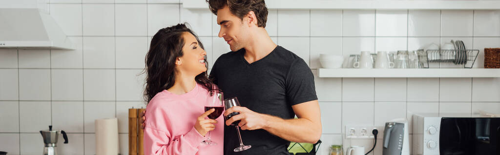 Panoramaaufnahme einer lächelnden Frau, die ihren Freund anschaut, während sie mit Wein in der Küche anstößt  - Foto, Bild