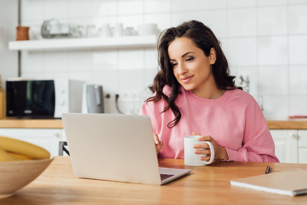 Επιλεκτική εστίαση της όμορφης γυναίκας κρατώντας φλιτζάνι καφέ κοντά στο φορητό υπολογιστή και σημειωματάριο στο τραπέζι της κουζίνας  - Φωτογραφία, εικόνα