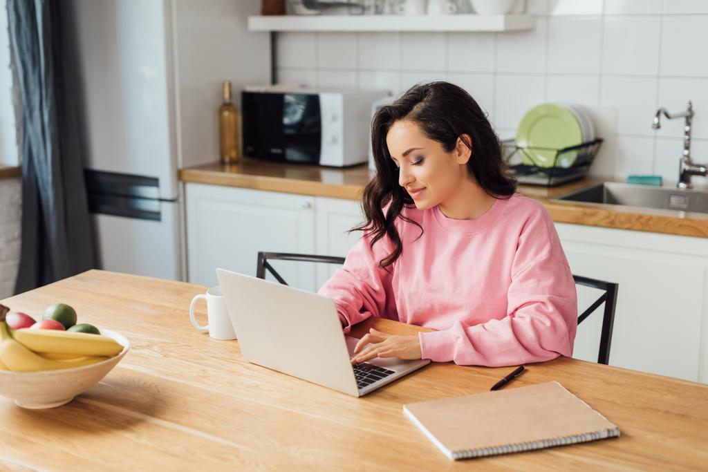 Επιλεκτική εστίαση των όμορφων freelancer χρησιμοποιώντας φορητό υπολογιστή κοντά στο σημειωματάριο και το φλιτζάνι του καφέ στην κουζίνα  - Φωτογραφία, εικόνα