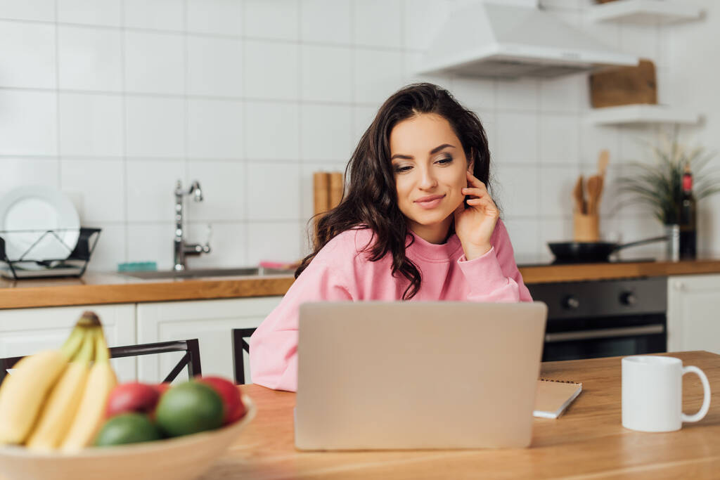 Selektywne skupienie atrakcyjnej freelancer patrząc na laptopa w pobliżu notebooka, filiżanki i świeże owoce w kuchni  - Zdjęcie, obraz