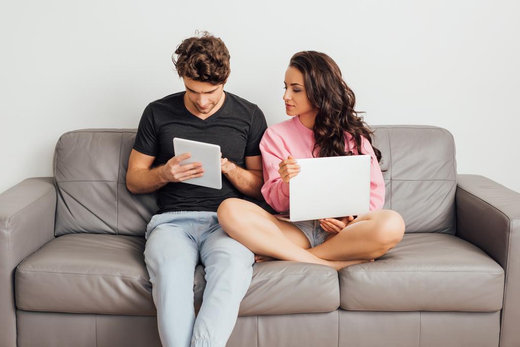 Junge Frau mit Laptop sitzt neben Freund mit digitalem Tablet auf Couch auf grauem Hintergrund  - Foto, Bild
