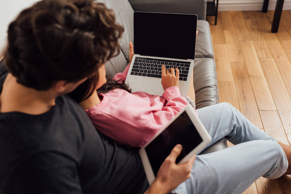 Enfoque selectivo del hombre sosteniendo la tableta digital mientras que la mujer utiliza el ordenador portátil con pantalla en blanco en el sofá en la sala de estar
 - Foto, imagen
