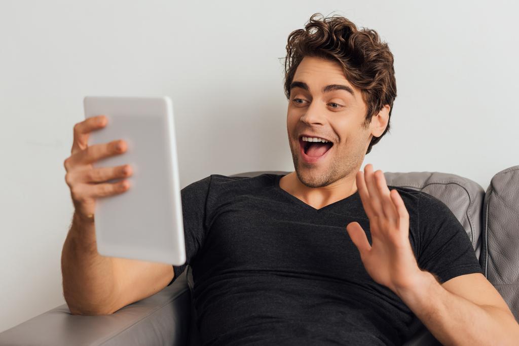 Foco seletivo de homem alegre acenando a mão enquanto tem chamada de vídeo no tablet digital no sofá no fundo cinza
  - Foto, Imagem