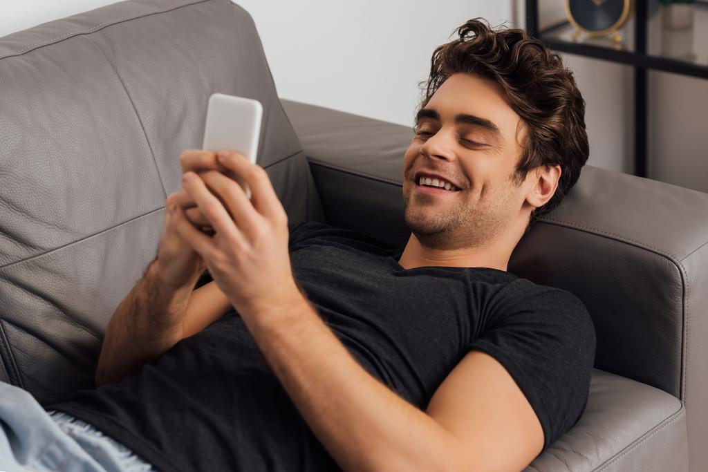 Focus selettivo dell'uomo sorridente che chiacchiera sullo smartphone mentre è sdraiato sul divano di casa  - Foto, immagini