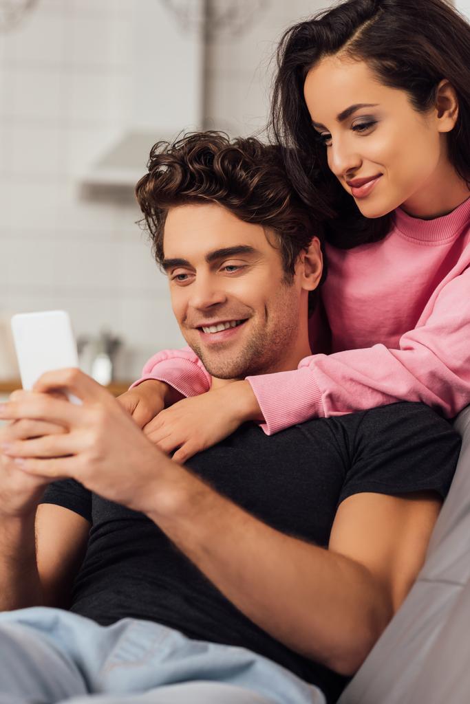 Enfoque selectivo de la hermosa mujer abrazando novio sonriente utilizando el teléfono inteligente en casa
  - Foto, imagen