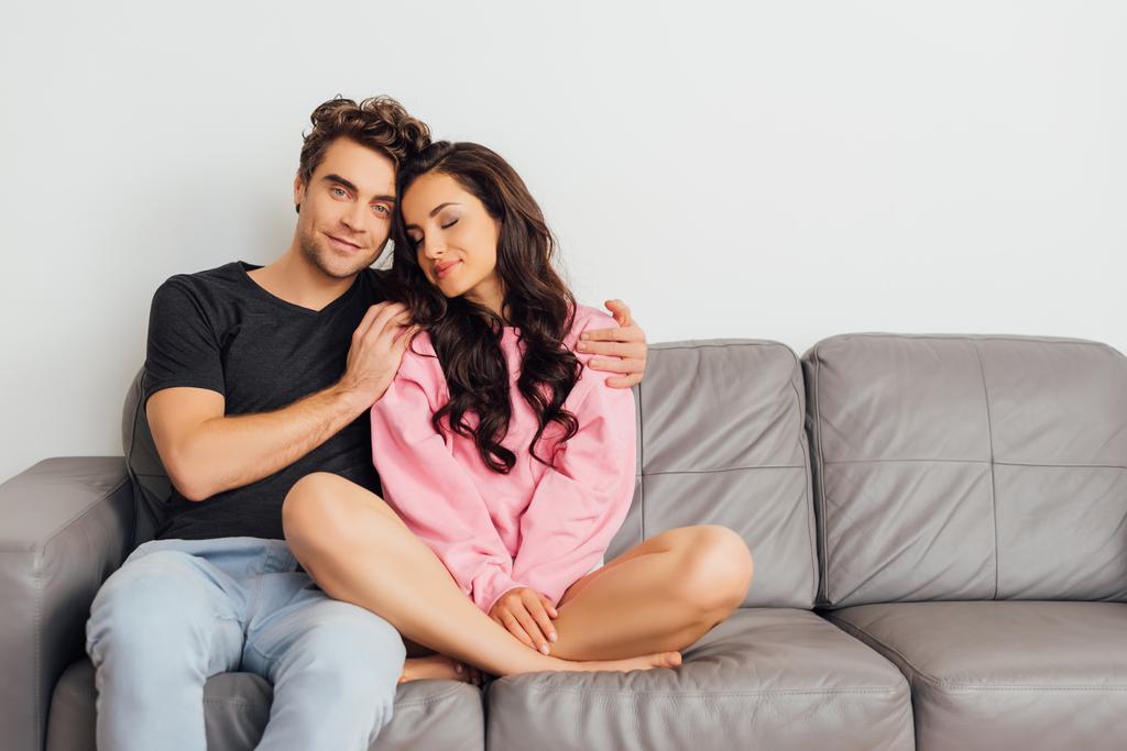 Hombre guapo abrazando a mujer hermosa con los ojos cerrados en sofá sobre fondo gris
  - Foto, Imagen