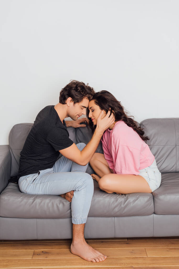 Boční pohled pohledný muž dotýká tvář krásné brunetky přítelkyně se zavřenýma očima na gauči  - Fotografie, Obrázek