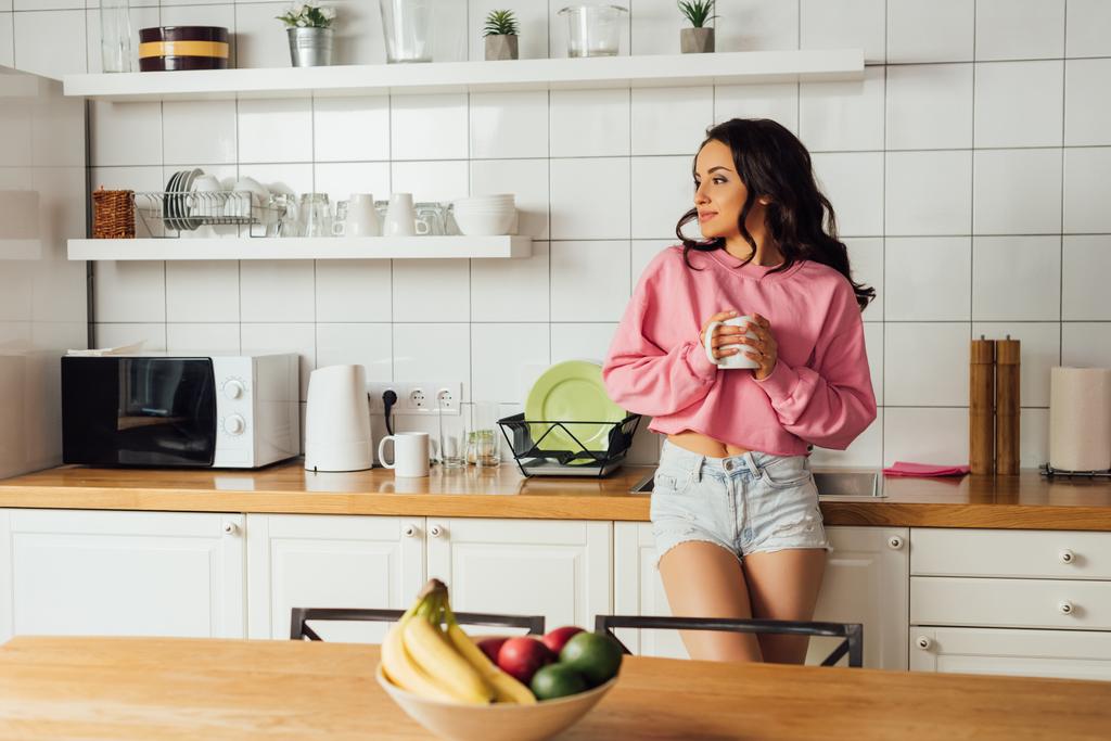 Selective focus of beautiful brunette woman holding cup of coffee in kitchen  - Photo, Image