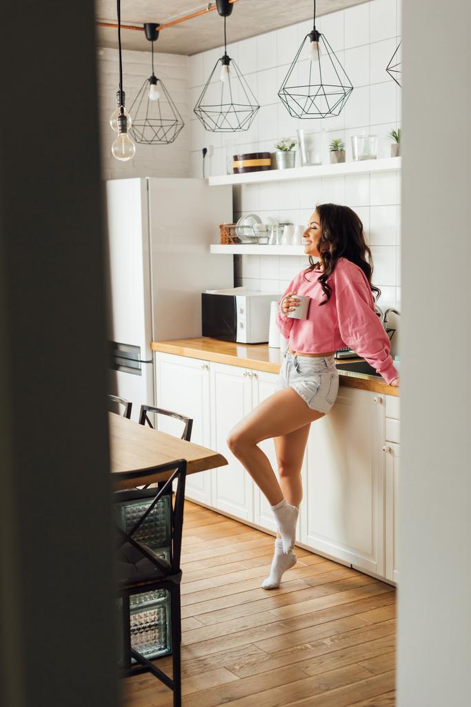 Επιλεκτική εστίαση του όμορφου χαμογελαστού κοριτσιού κρατώντας φλιτζάνι καφέ στην κουζίνα  - Φωτογραφία, εικόνα