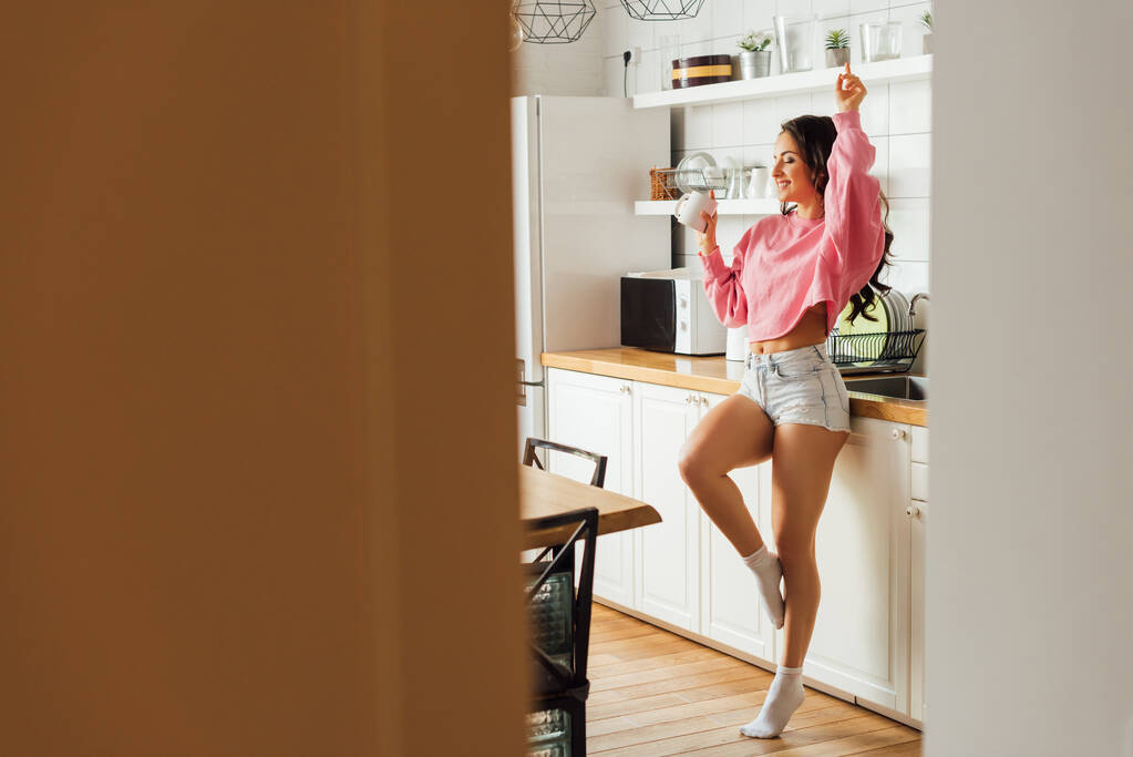 Selective focus of cheerful woman holding cup of coffee in kitchen at morning  - Photo, Image