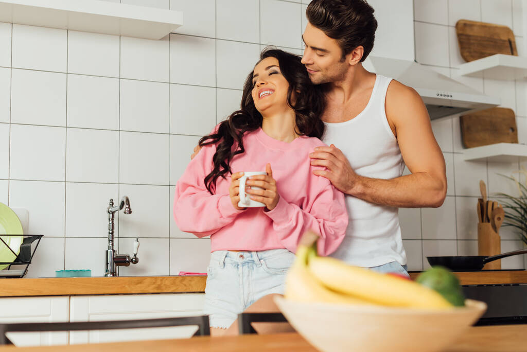 Selektywne skupienie człowieka obejmującego piękną uśmiechniętą dziewczynę z filiżanką kawy w kuchni  - Zdjęcie, obraz