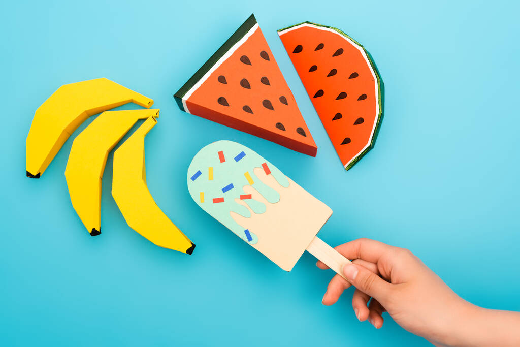 cropped view of woman holding paper ice cream with sprinkles near watermelon, bananas on blue background - Photo, Image
