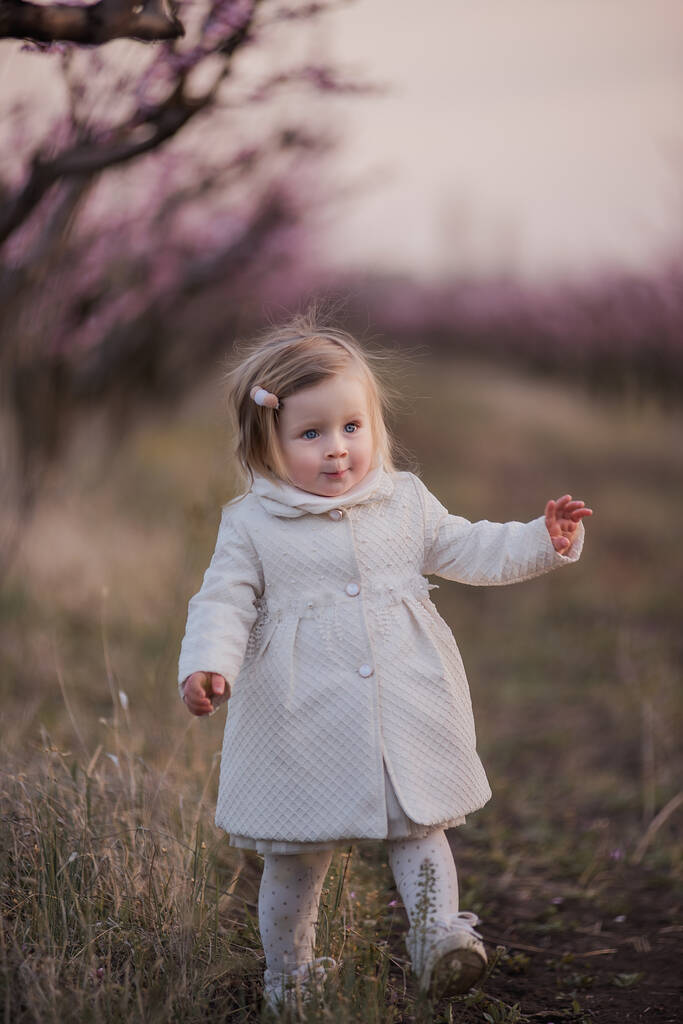 Портрет маленького красивого ребенка с голубыми глазами в белом халате, который ходит на пикник со своими родителями в цветущих садах на закате. Путешествует с маленькими детьми. Модная блондинка. - Фото, изображение