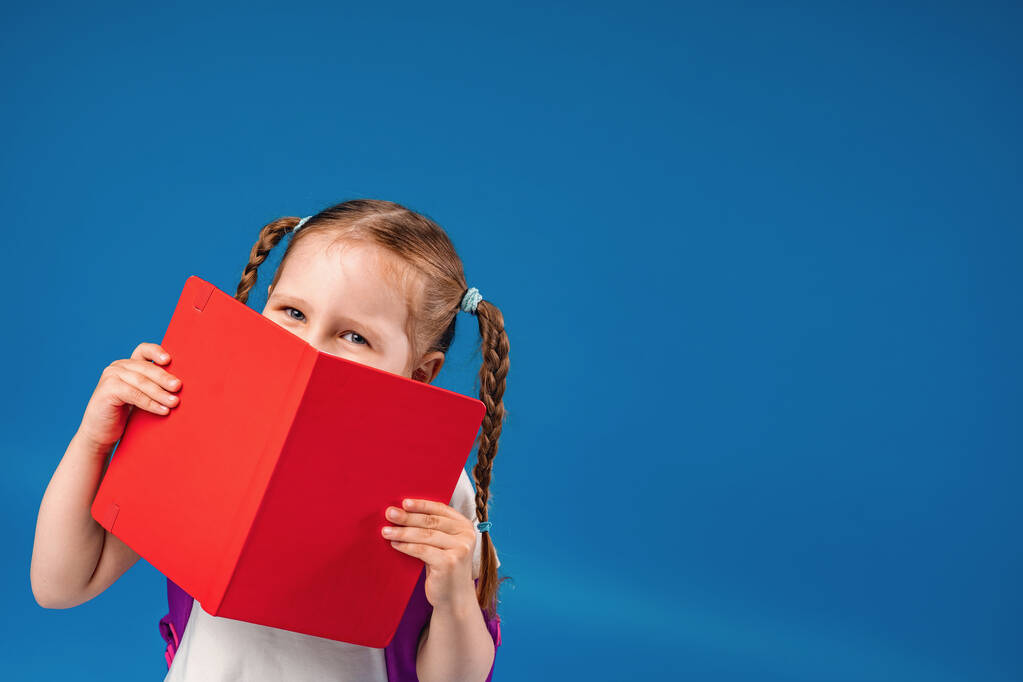 elbűvölő csintalan kislány szemüveggel mosolyog a kamerába, és elbújik egy könyv mögé, szórakozás közben iskolai osztályok kék háttérrel. Vissza az iskolába.. - Fotó, kép