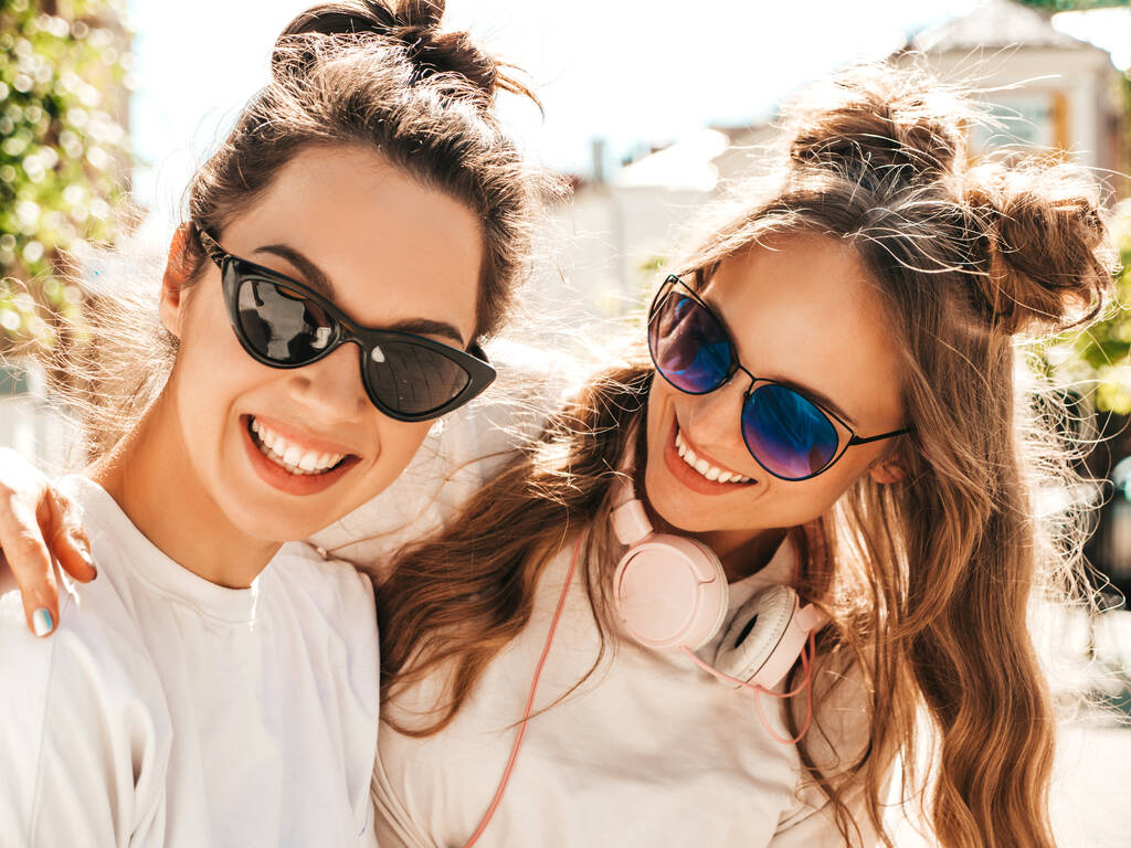 Portrait von zwei jungen schönen lächelnden Hipster-Frauen in trendigen sommerweißen T-Shirt-Kleidern. Sexy unbeschwerte Frauen posieren vor Straßenhintergrund. Positive Models, die Spaß haben, sich umarmen und durchdrehen - Foto, Bild
