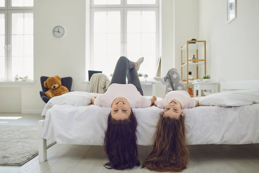Mãe alegre e filha com cabelo bonito deitado de cabeça para baixo na cama em casa. Pais e criança se divertindo no quarto
 - Foto, Imagem