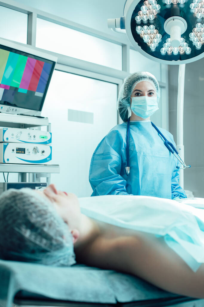 Пациент на медицинском диване в операционной. Молодая женщина-врач со стетоскопом на шее, стоящая рядом с ним - Фото, изображение