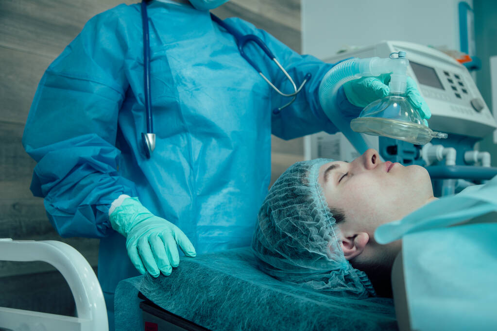 Jovem adulto deitado em uma mesa médica na sala de cirurgia e um médico profissional segurando uma máscara anestésica acima de seu rosto
 - Foto, Imagem