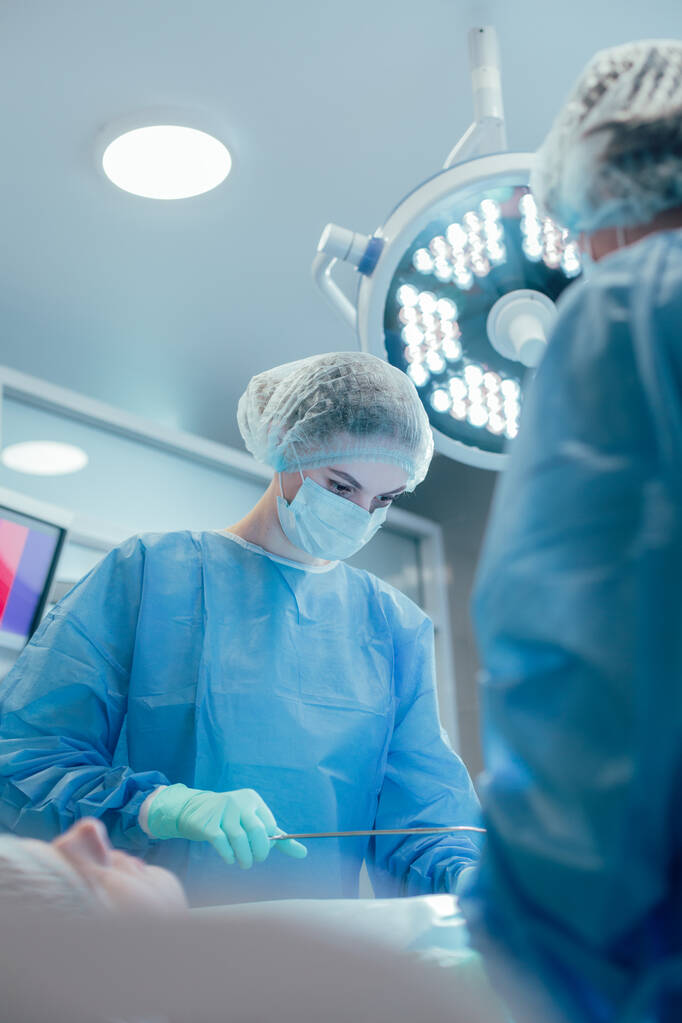 Профессиональная медицинская работница, выполняющая хирургическую операцию вместе со своими коллегами и с помощью щипцов - Фото, изображение