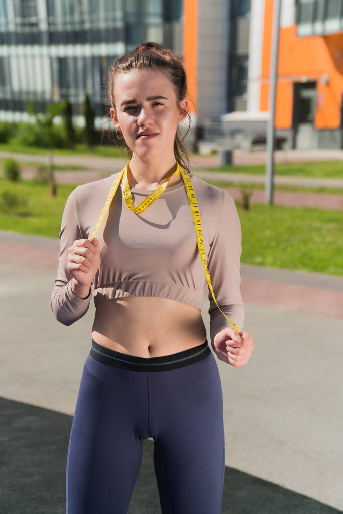 Smutná štíhlá dívka drží v rukou centimetrovou pásku. Koncept nevhodného hubnutí žena fitness trenér v parku - Fotografie, Obrázek