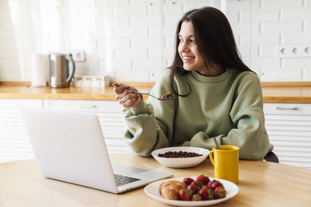 Foto de mulher bonita alegre usando laptop enquanto toma café da manhã na cozinha moderna
 - Foto, Imagem