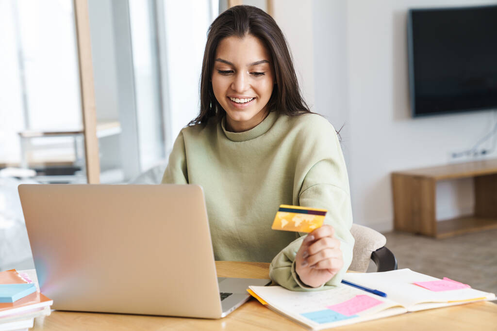 Fotografie veselá hezká studentka držící kreditní kartu a pomocí notebooku při domácím úkolu v obývacím pokoji - Fotografie, Obrázek
