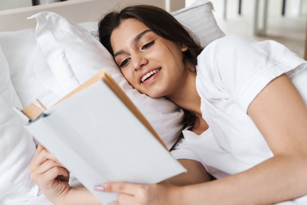 Zdjęcie skupionej uśmiechniętej pięknej kobiety czytającej książkę leżącej na łóżku po śnie lub drzemce - Zdjęcie, obraz
