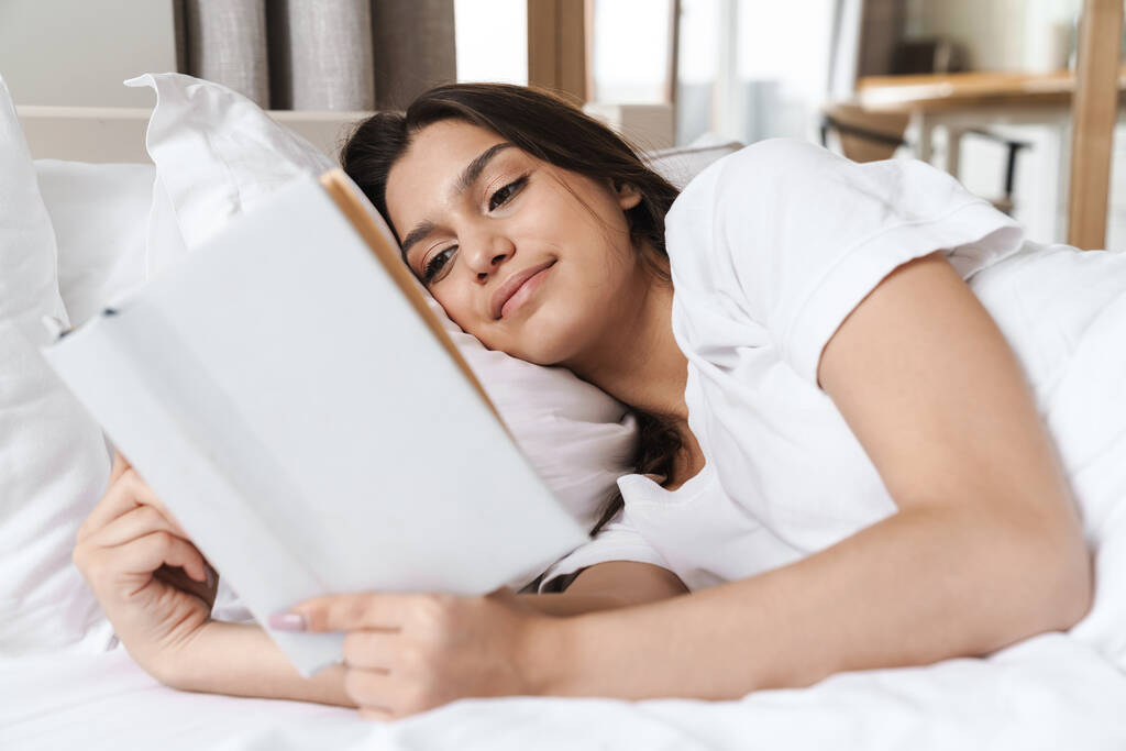 Foto de mulher muito sorridente lendo livro enquanto estava deitado na cama no travesseiro branco após o sono ou cochilo
 - Foto, Imagem