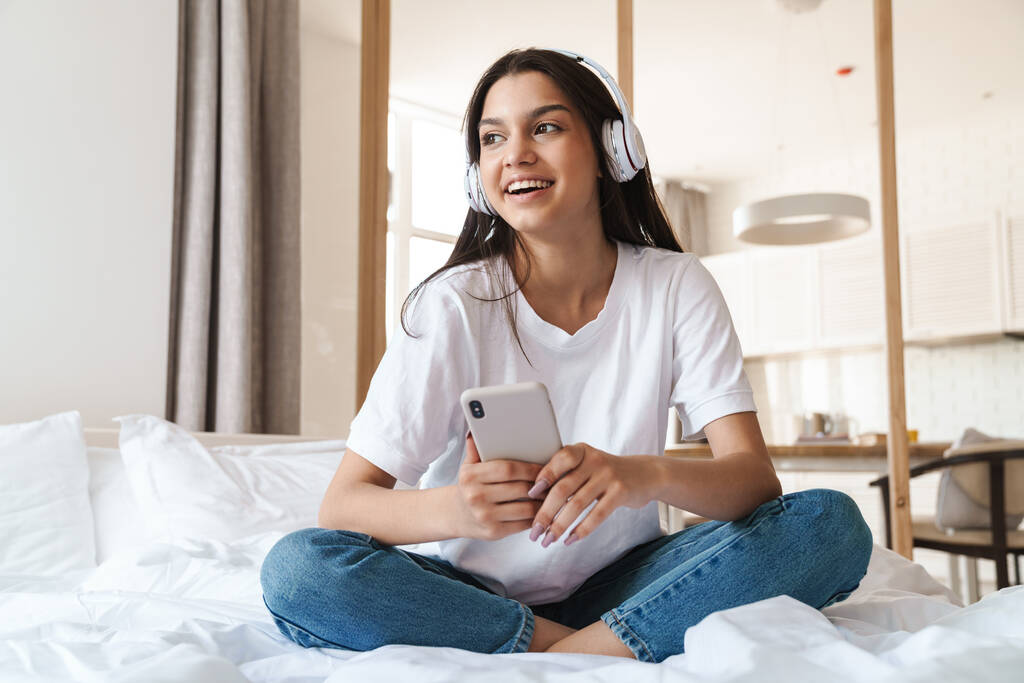 Foto de una hermosa mujer sonriendo mientras usa auriculares inalámbricos y teléfonos celulares en la cama en casa
 - Foto, imagen