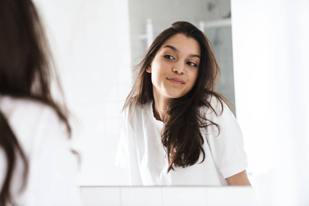 Fotografie veselé pěkné ženy s brunetkou vlasy usmívat při pohledu na zrcadlo v koupelně - Fotografie, Obrázek