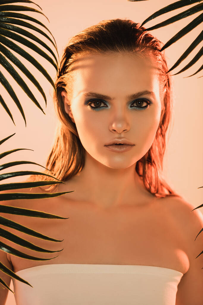 beautiful woman with makeup looking at camera near palm leaves on beige - Photo, Image