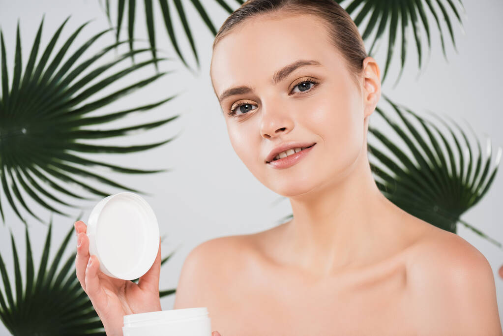 happy woman holding container with face cream near palm leaves on white - Photo, Image