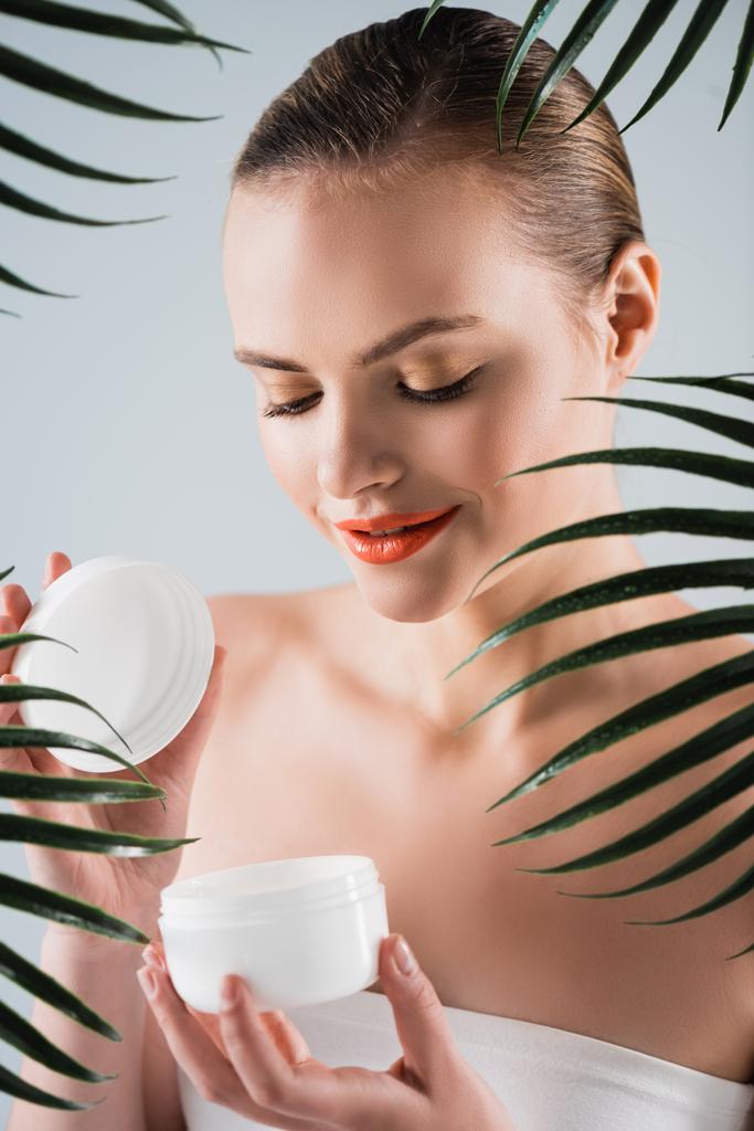 happy girl with makeup holding container with face cream near palm leaves on white - Photo, Image