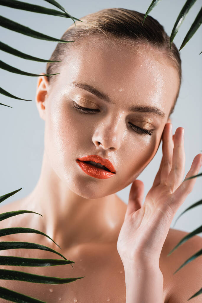 atrakcyjna i mokra kobieta z makijażem dotykająca twarzy w pobliżu liści palmy na białym - Zdjęcie, obraz