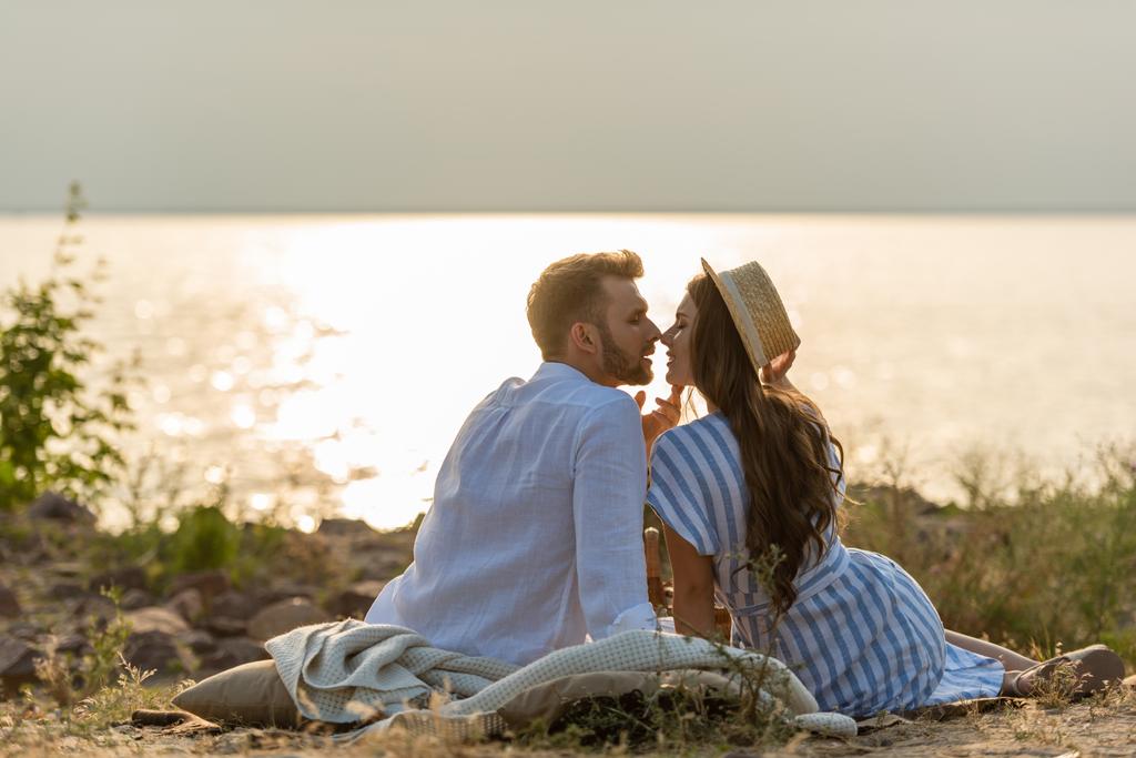 vista lateral de homem bonito e mulher atraente em chapéu de palha sentado perto do lago
  - Foto, Imagem