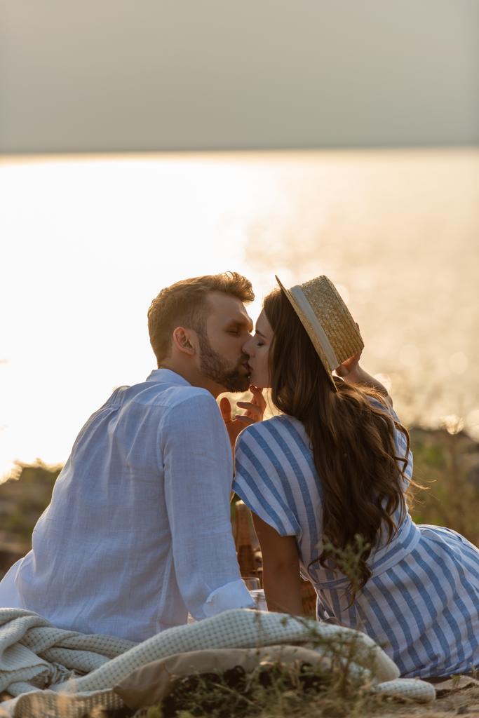 Hasır şapkalı yakışıklı adam ve çekici kadının göl kenarında öpüşmelerinin yan görüntüsü.  - Fotoğraf, Görsel