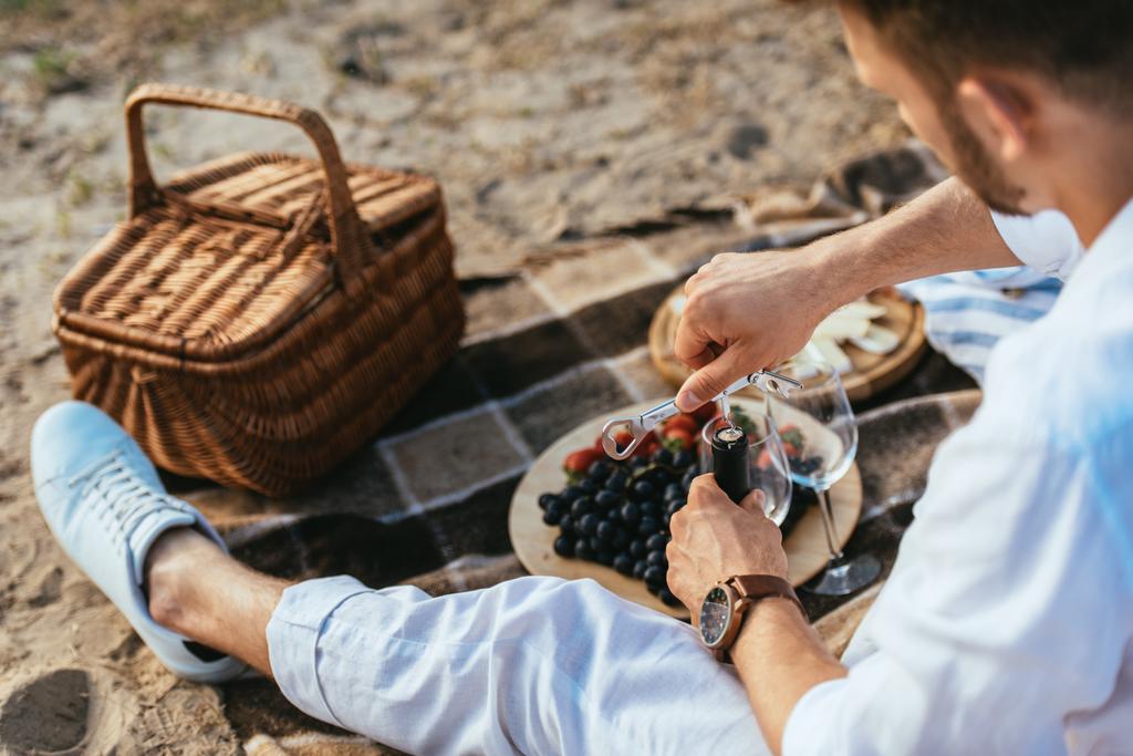 szelektív fókusz szakállas ember nyitó üveg bor közelében gyümölcsök tányéron és fonott kosár takarón  - Fotó, kép