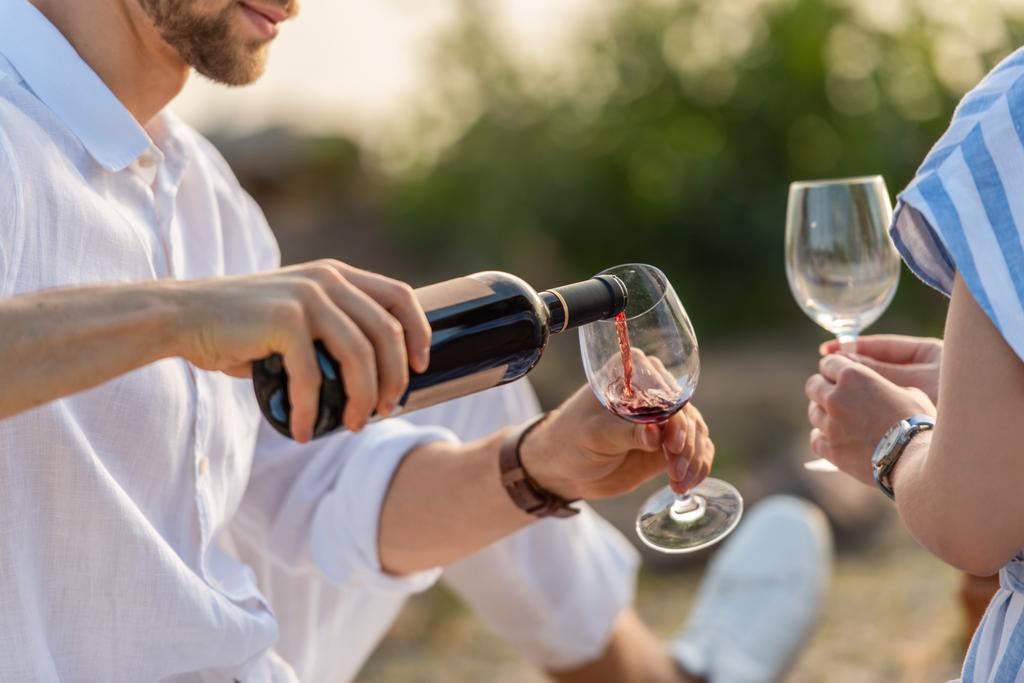 περικοπή άποψη του γενειοφόρου άνδρα κρατώντας μπουκάλι, ενώ ρίχνει κόκκινο κρασί σε ποτήρι κοντά γυναίκα - Φωτογραφία, εικόνα