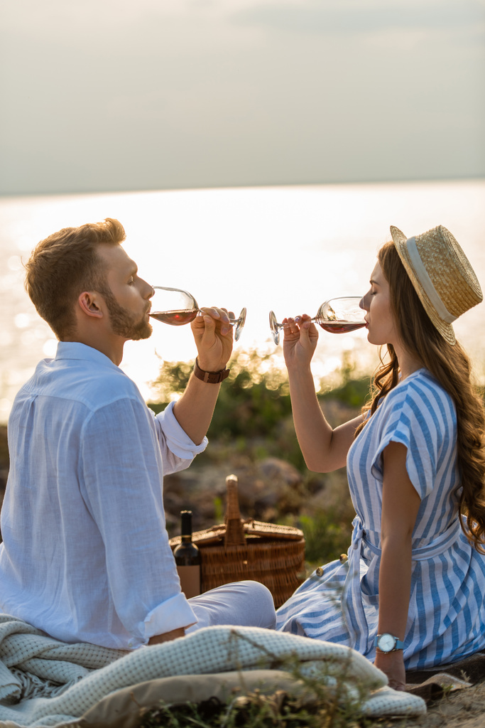 vista lateral do casal segurando óculos enquanto bebe vinho tinto perto do lago
  - Foto, Imagem