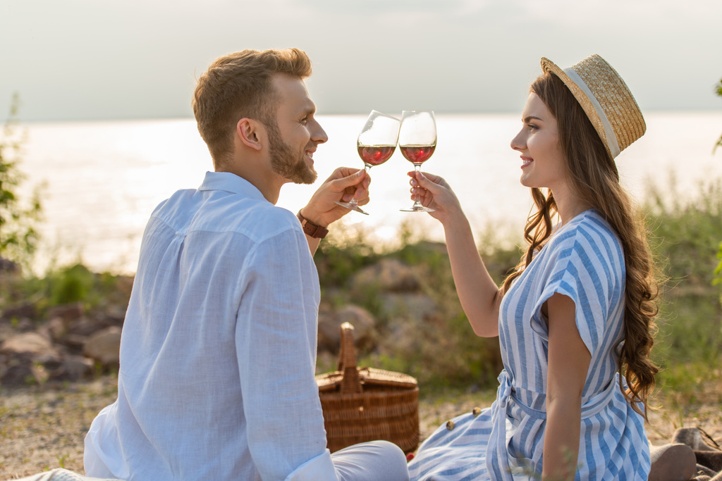vista lateral de alegre pareja tintineo vasos con vino tinto mientras está sentado cerca del lago
  - Foto, imagen