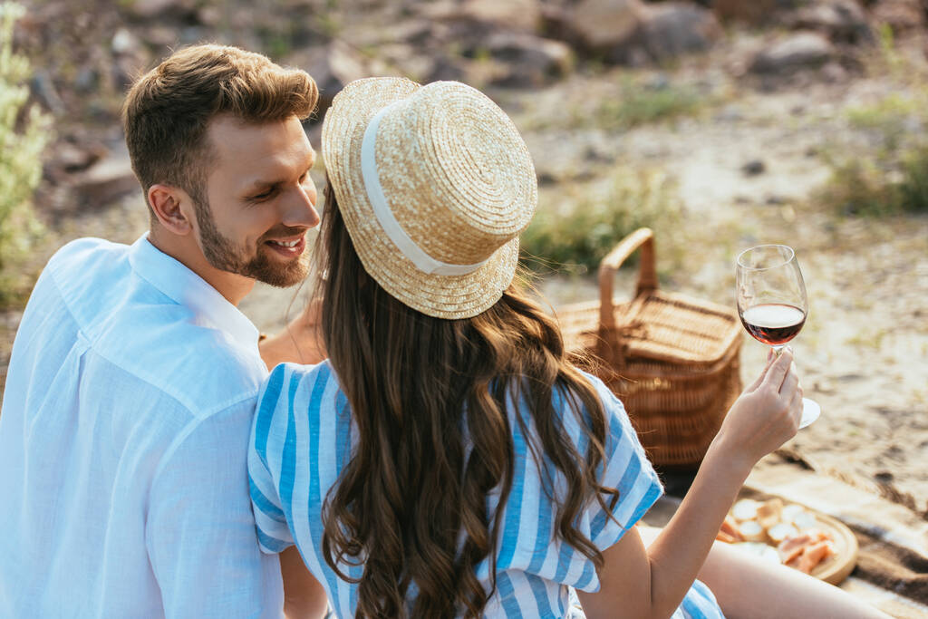 селективное внимание девушки в соломенной шляпе держа стакан с красным вином рядом счастливый парень  - Фото, изображение