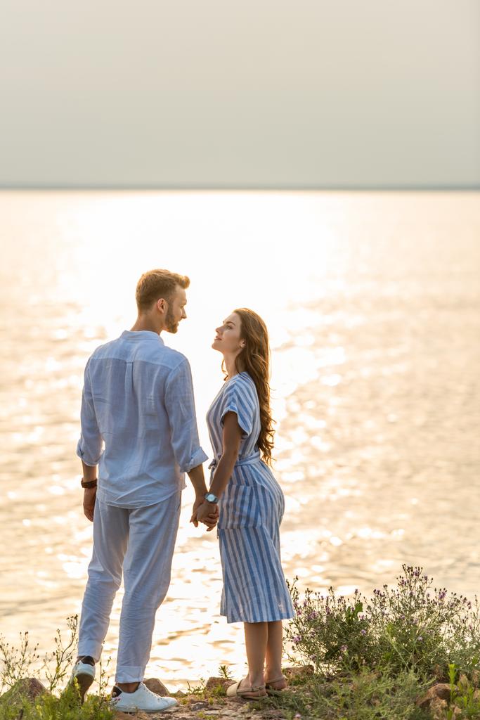 Красивый мужчина и привлекательная женщина держатся за руки возле озера  - Фото, изображение
